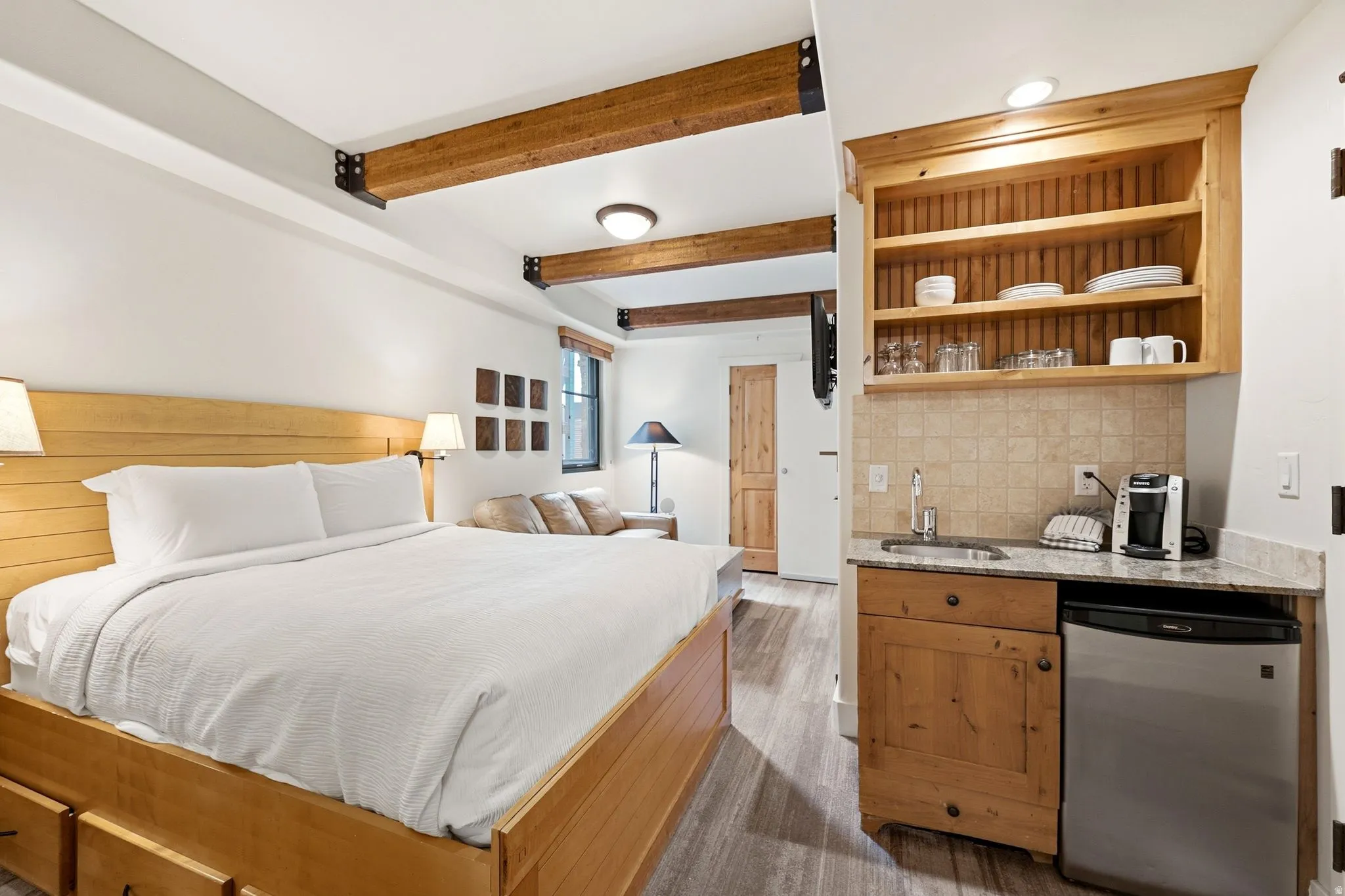 Exposed wood beams, wood-finish flooring, built-in kitchenette with a compact refrigerator, light-toned wood cabinetry, and a stone-finish countertop
