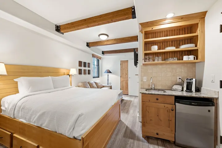 Exposed wood beams, wood-finish flooring, built-in kitchenette with a compact refrigerator, light-toned wood cabinetry, and a stone-finish countertop