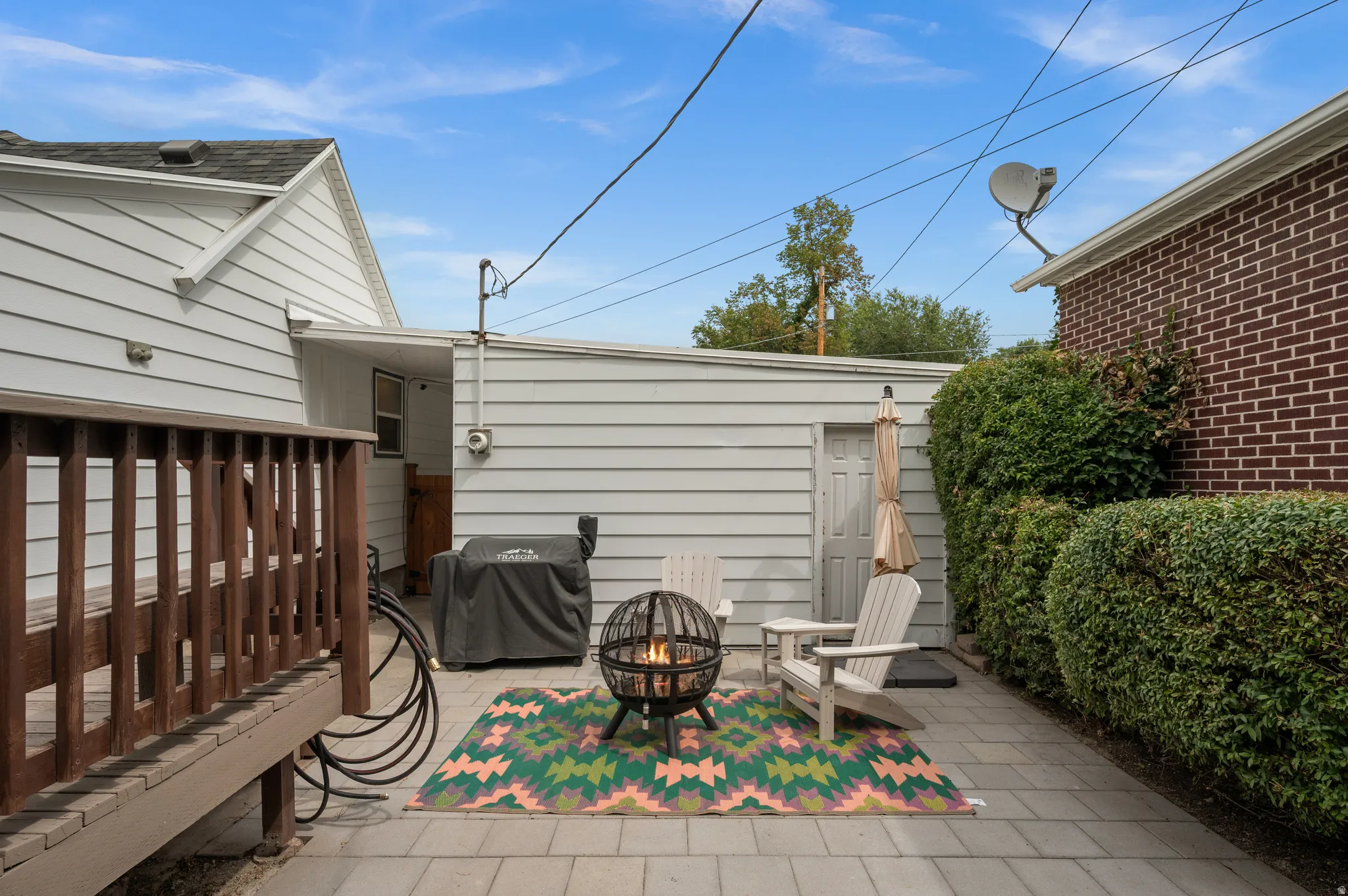 Paved patio area, wood deck, building with horizontal siding, brick exterior wall, and manicured shrubs
