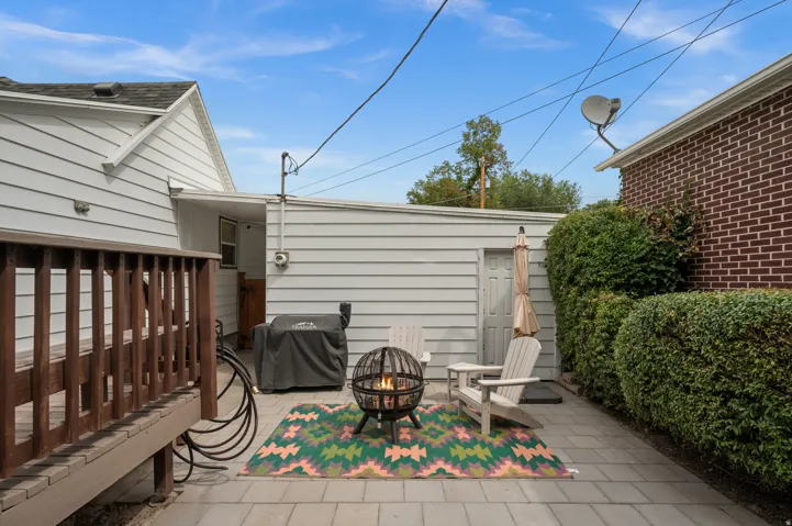 Paved patio area, wood deck, building with horizontal siding, brick exterior wall, and manicured shrubs