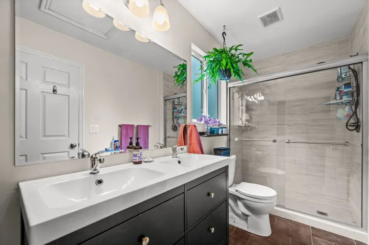 Dual sink vanity with dark wood-finish cabinetry, chrome fixtures, large wall mirror, walk-in shower with sliding glass door, and tiled shower surround