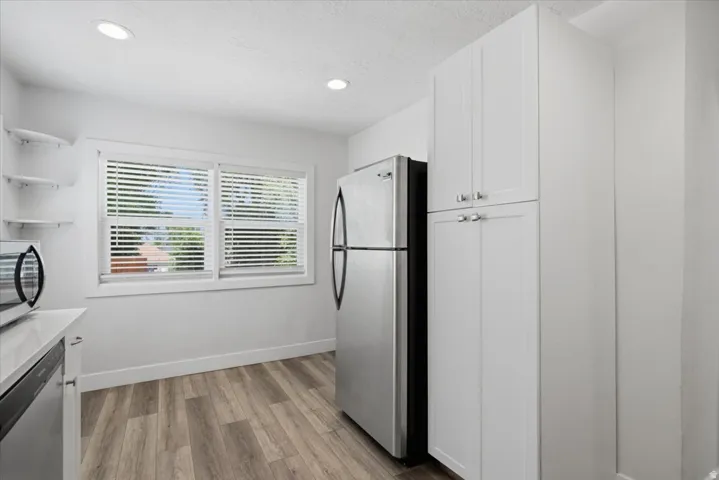 White shaker-style cabinetry with silver hardware, stainless steel refrigerator, recessed lighting, wood-finish flooring, and a large window with blinds