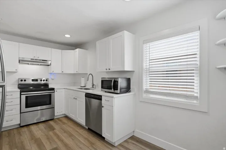 Contemporary kitchen featuring white cabinetry, stainless steel appliances, light countertops, a window with blinds, and wood-finish flooring