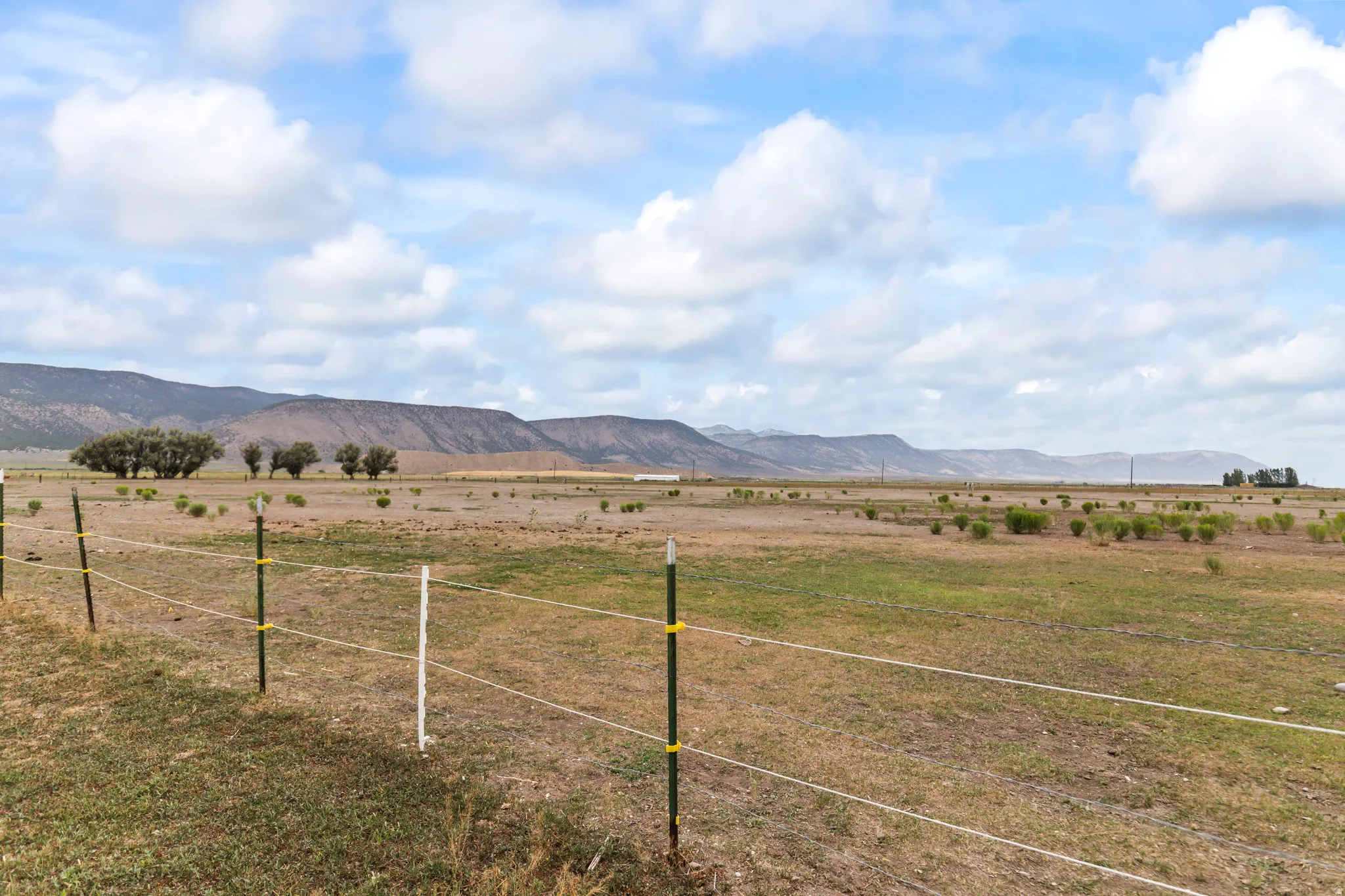Expansive rural landscape featuring a distant mountain range, open fields with scattered vegetation, and a clear sky with clouds