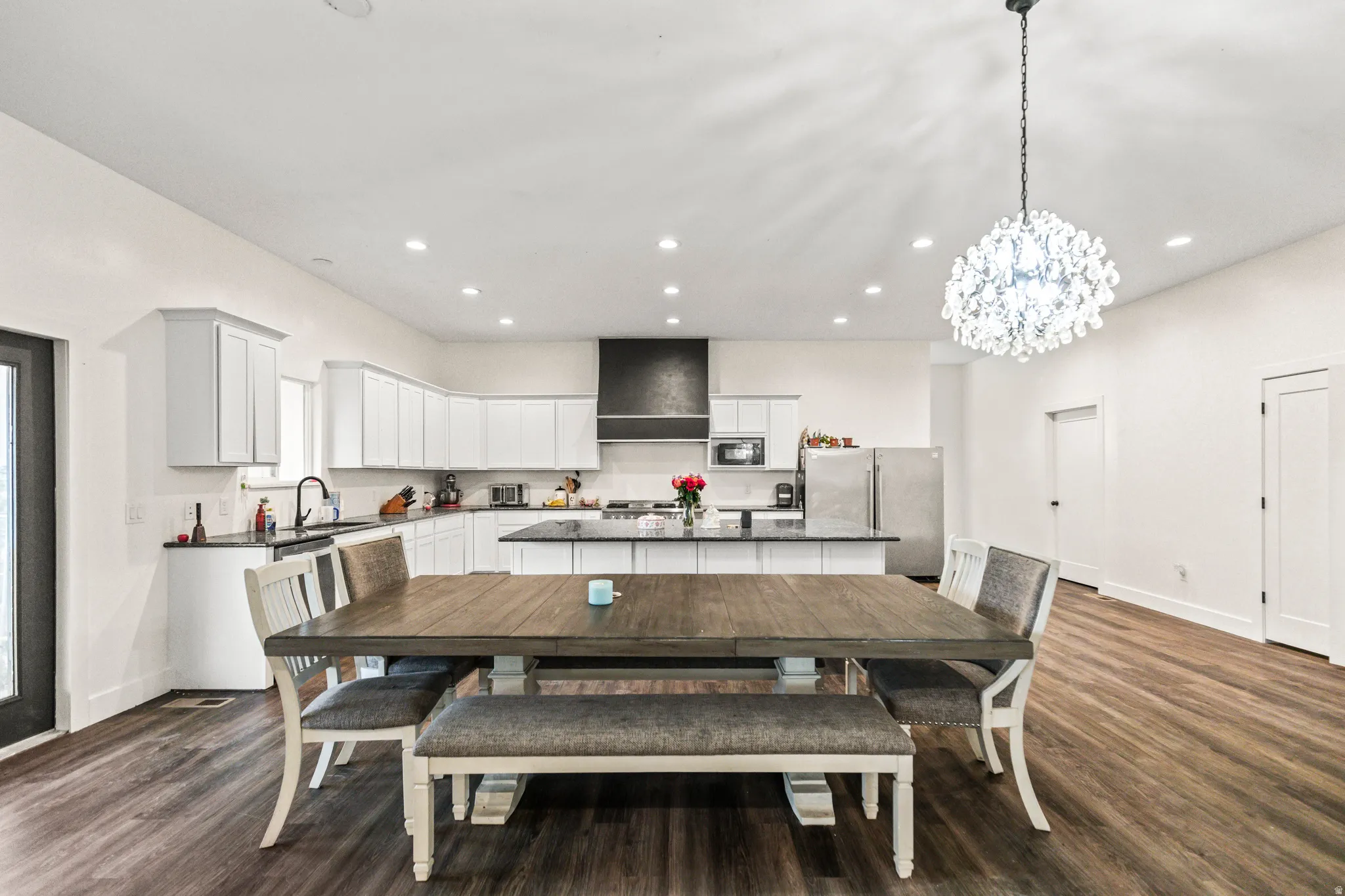 Open-concept kitchen and dining area featuring wood-finish flooring, white cabinetry, a kitchen island with dark countertops, a dark range hood, and a decorative chandelier