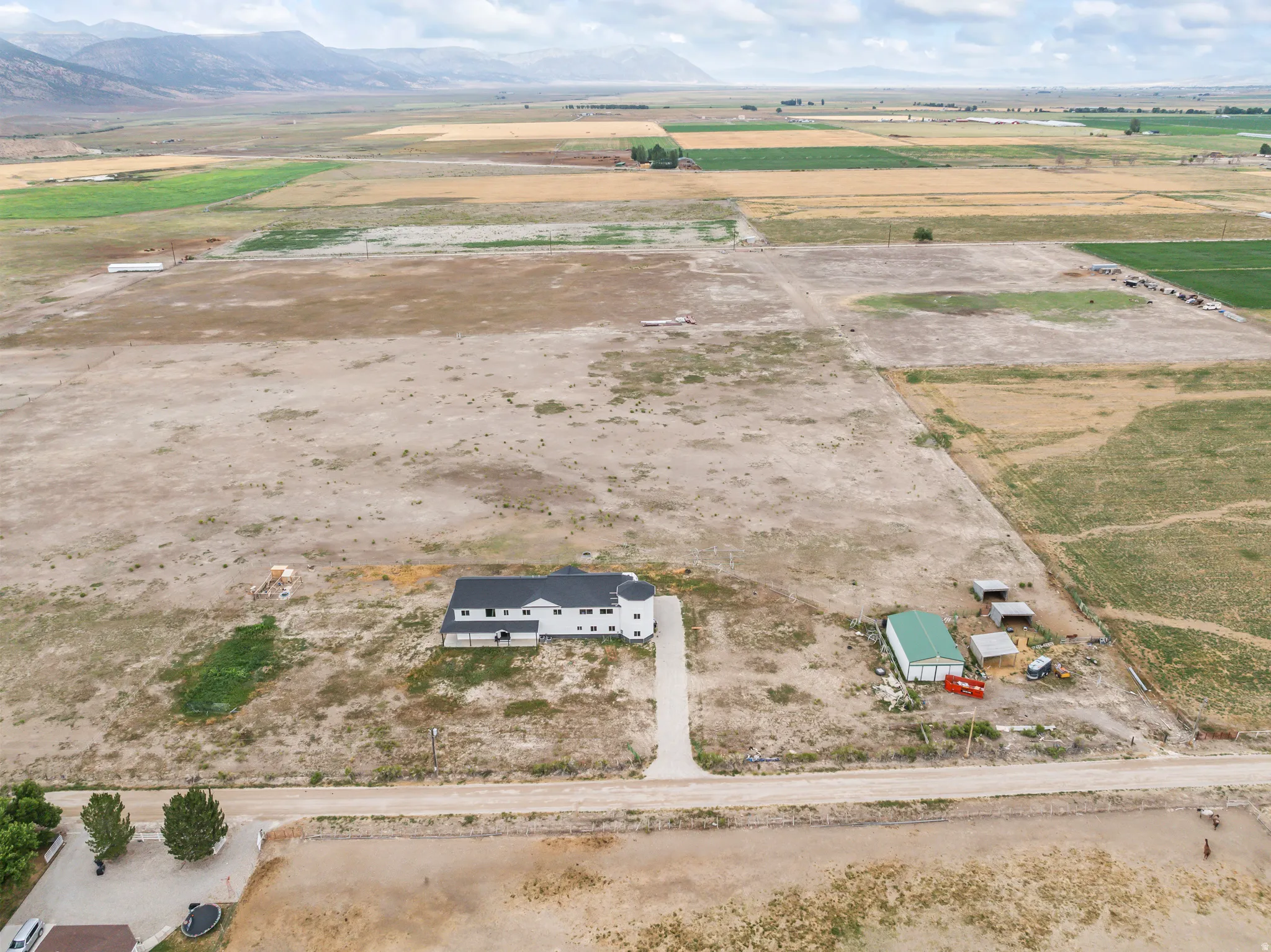 Expansive property featuring a white residence with a black roof, a detached green outbuilding, a gravel driveway, and surrounding agricultural land with distant mountains