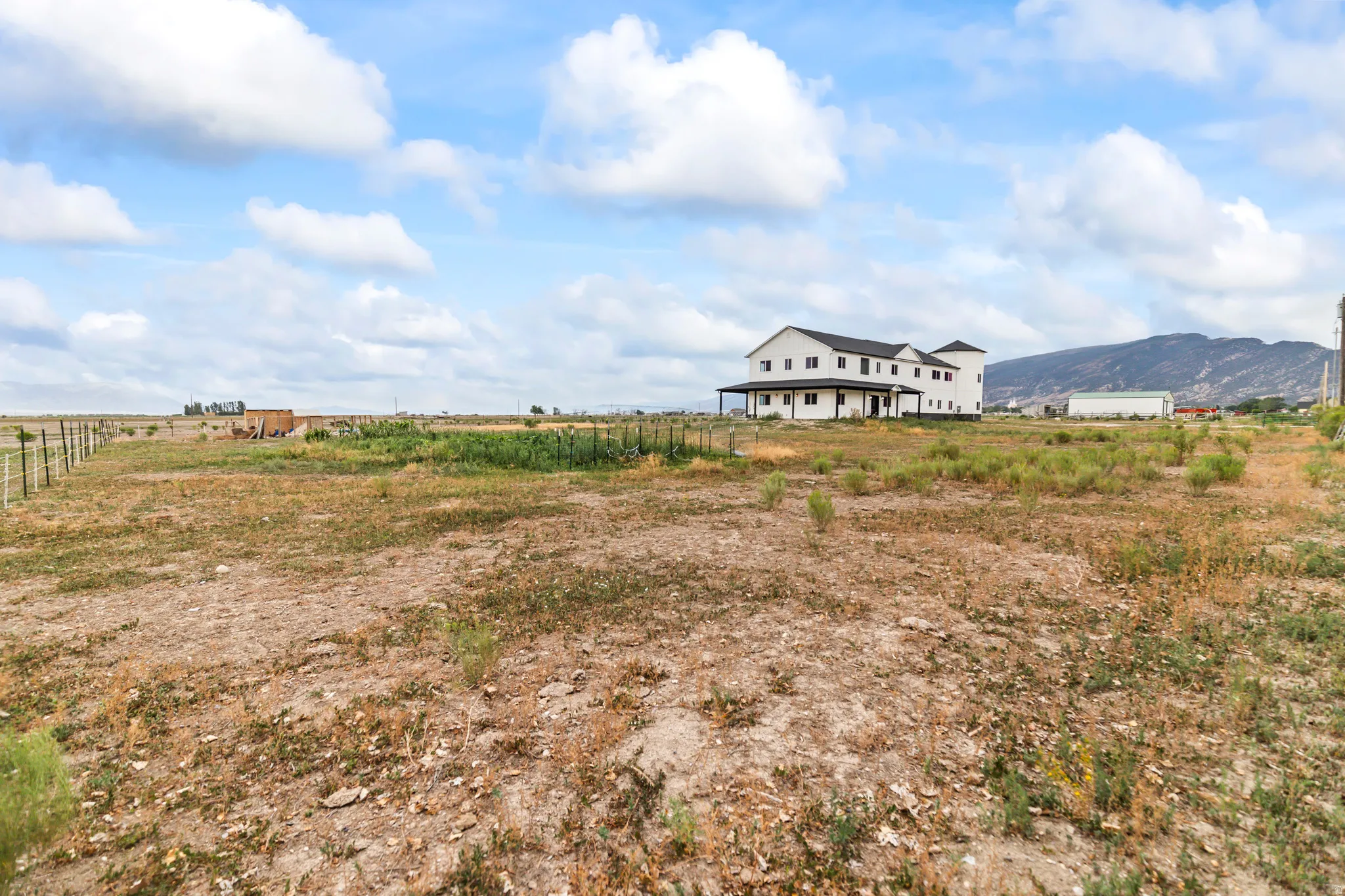 Expansive open land with a distant two-story property featuring a wrap-around porch and a dark roof