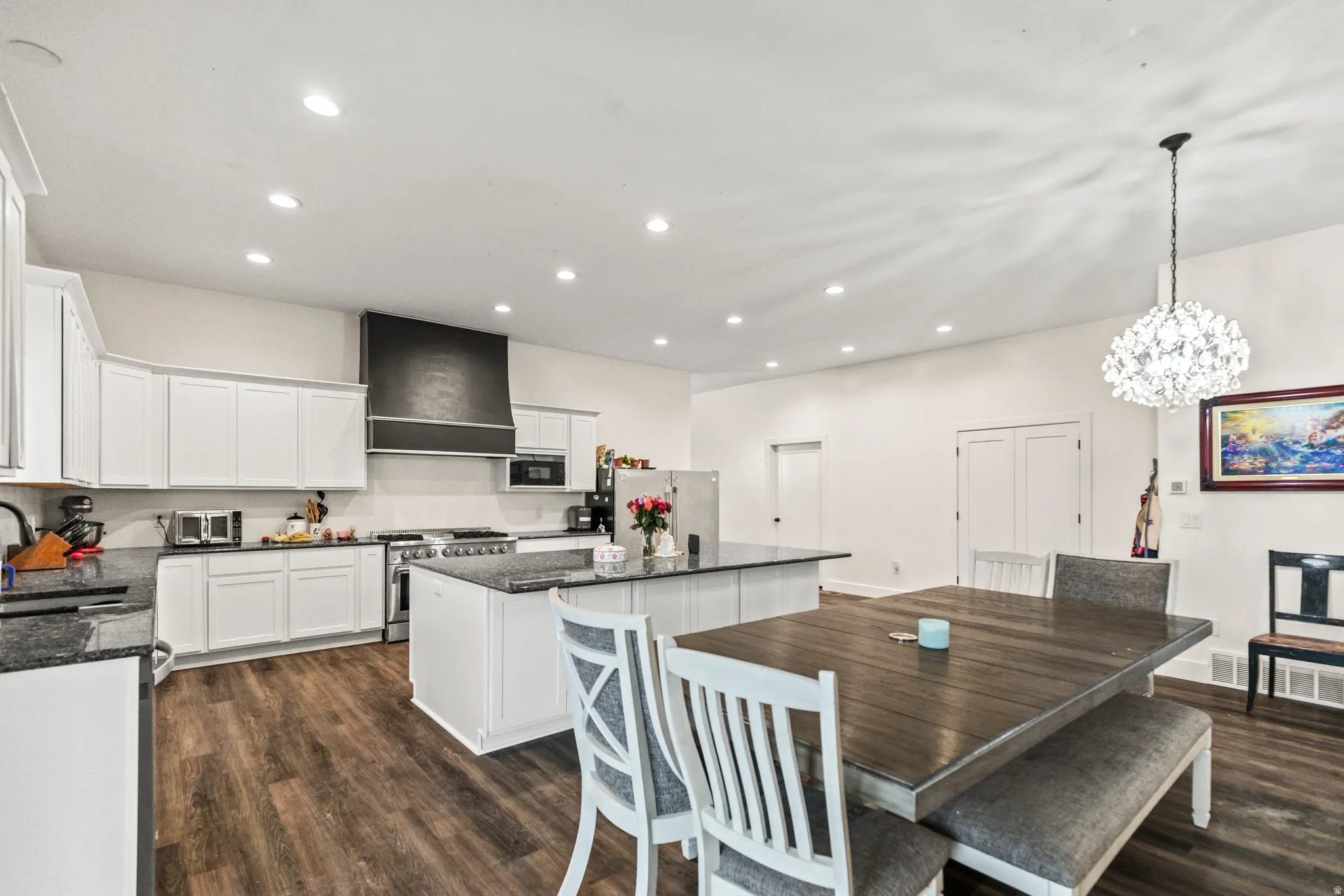 White cabinetry, dark stone countertops, wood-finish flooring, recessed lighting, and a crystal chandelier
