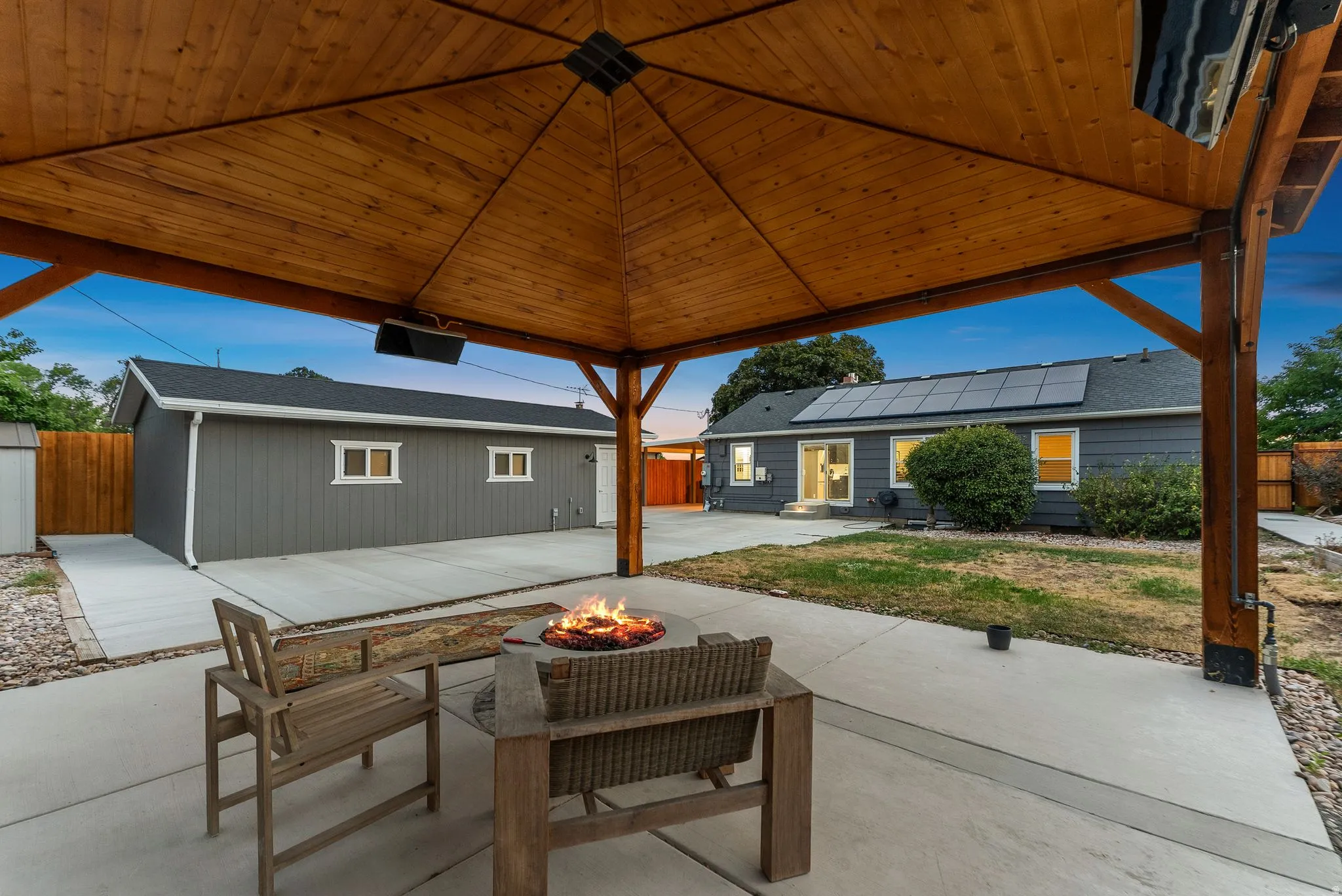 Expansive concrete patio with a wood-framed gazebo, solar panels installed on the roof, a detached garage, and a fire pit