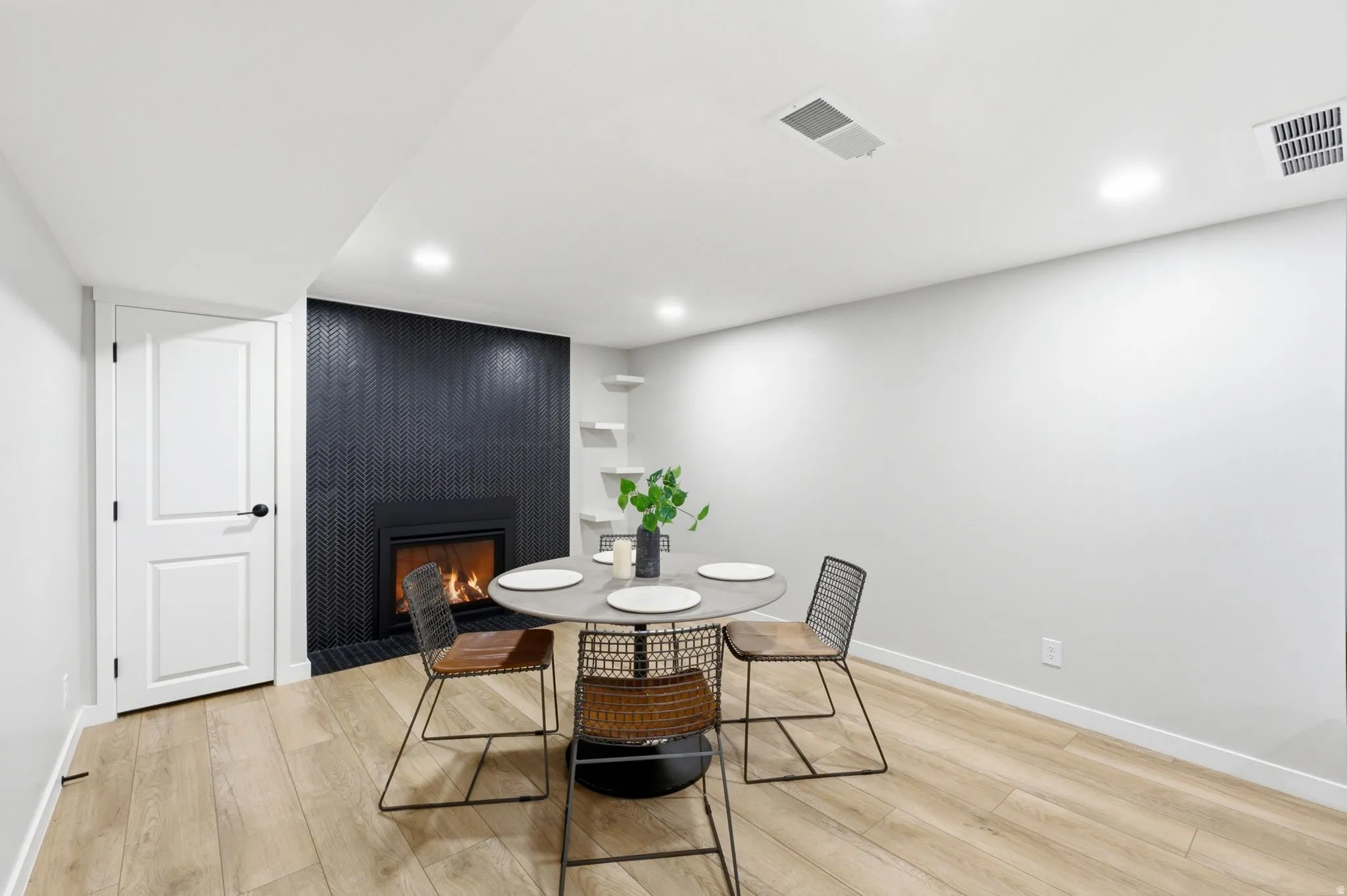 Modern recreation room featuring warm wood-finish flooring, a round accent table with geometric metal base, and a striking black vertical tile fireplace with active firebox