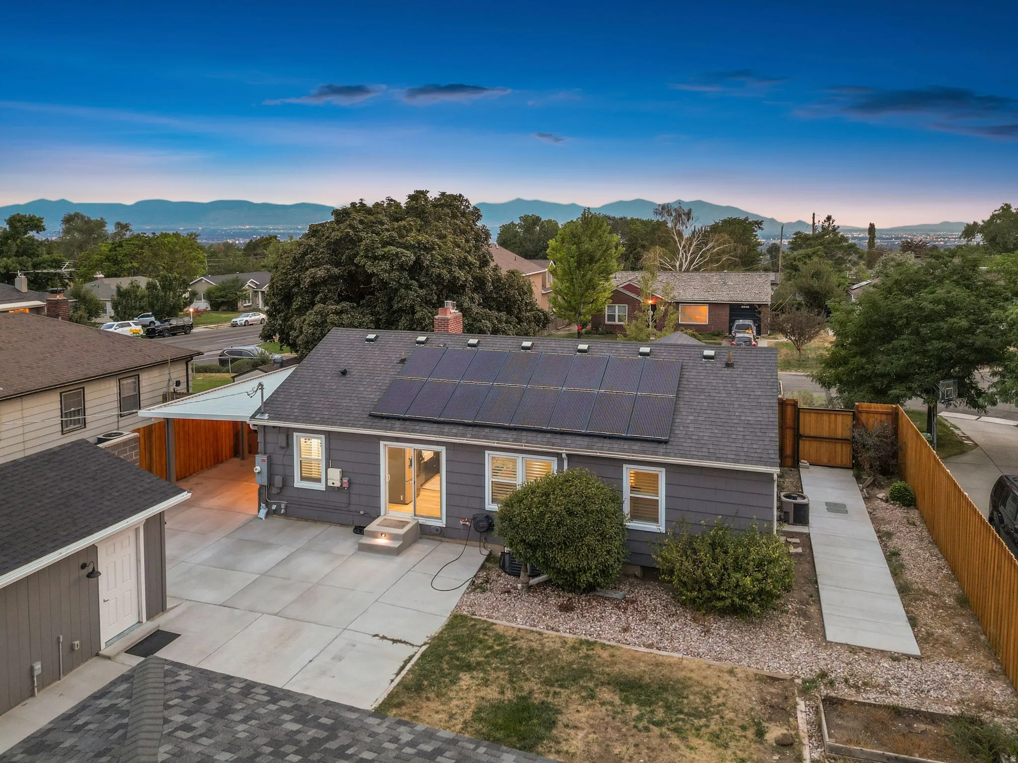 Modern residence featuring a dark composite roof with solar panels, expansive concrete patio with integrated landscaping, and mature specimen trees throughout the grounds