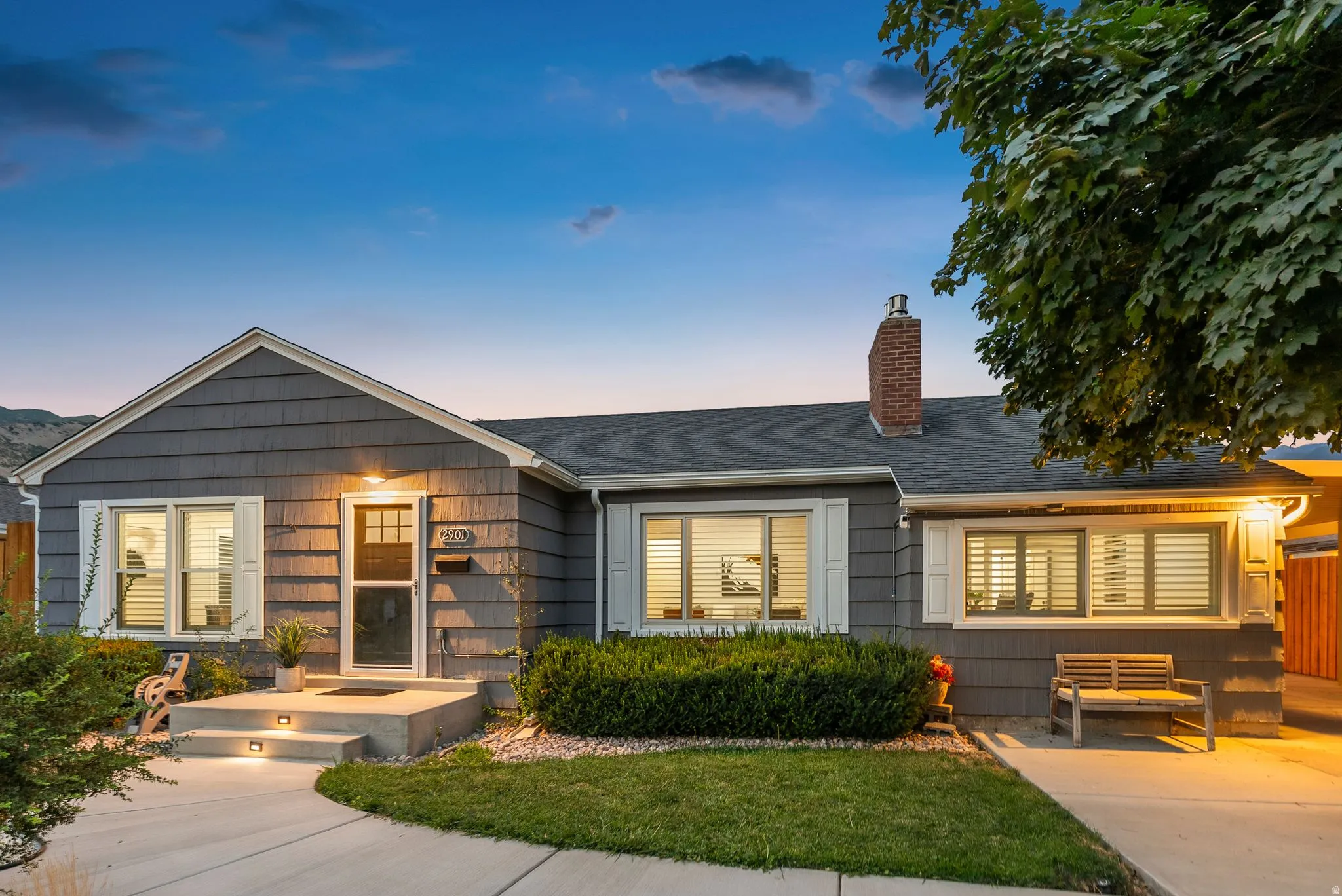 Modern farmhouse residence featuring a charming front facade with board-and-batten siding, welcoming covered entry porch with stepped concrete approach, large multi-pane windows flooding interior spaces with natural light, brick chimney detail, and manicu