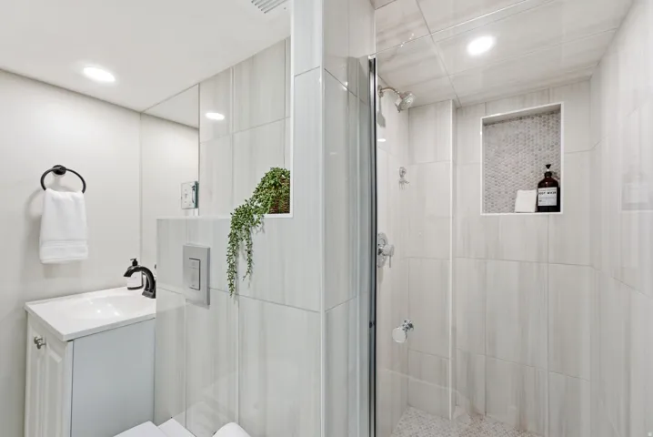 White vanity with a single basin sink and matte black faucet, glass-enclosed shower, light-toned tiled walls, recessed shower niche with mosaic tile backing, and polished chrome shower fixtures