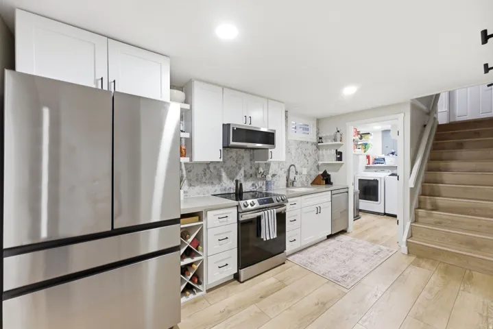 Stainless steel appliances, white cabinetry with dark hardware, stone-finish backsplash, wood-finish flooring, and a built-in wine rack