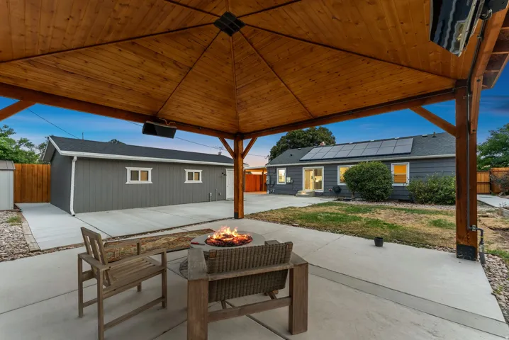 Expansive concrete patio with a wood-framed gazebo, solar panels installed on the roof, a detached garage, and a fire pit
