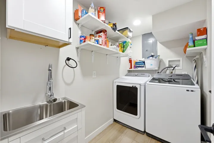 Efficient laundry room featuring side-by-side white front-load washer and dryer, stainless steel utility sink with chrome faucet, wood-finish flooring, white cabinetry with natural wood accents, and wall-mounted shelving with ample storage for household s