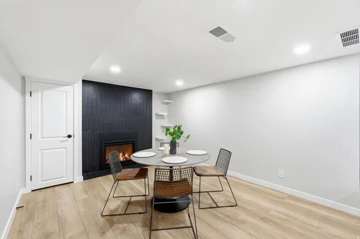 Modern recreation room featuring warm wood-finish flooring, a round accent table with geometric metal base, and a striking black vertical tile fireplace with active firebox