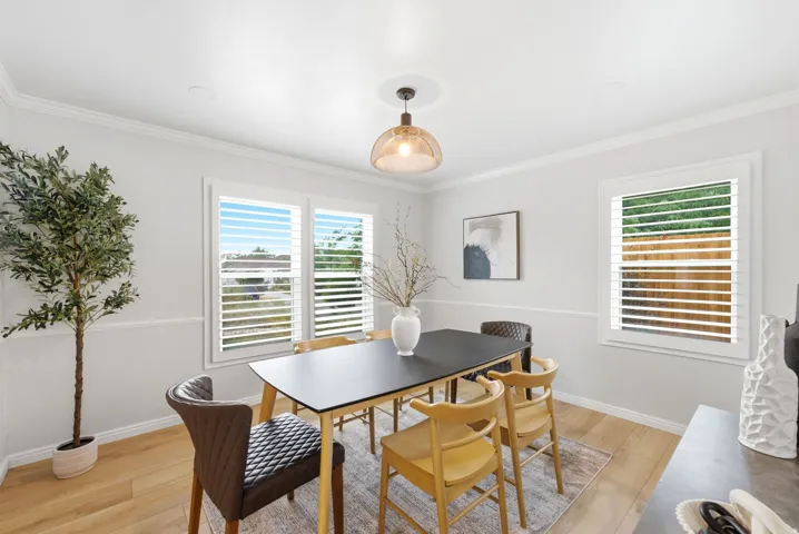Bright dining room featuring wood-finish flooring, a substantial dining table with seating for six, and abundant natural light from dual multi-pane windows with plantation shutters