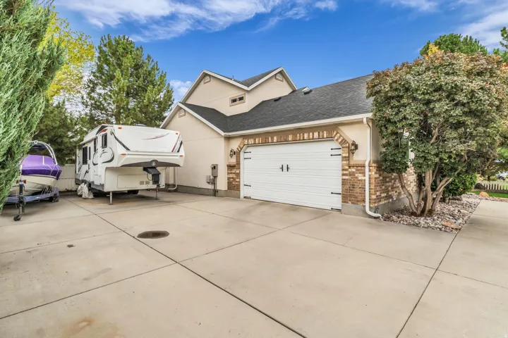 Expansive concrete driveway, attached garage with white door and decorative hardware, brick detailing on garage facade, exterior lighting fixture, and established landscaping