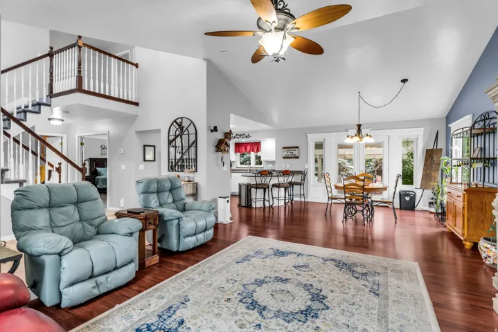 Open-concept living area featuring vaulted ceilings, wood-finish flooring, a white spindle staircase with dark wood handrails, a kitchen with a breakfast bar, and a dining area with multiple windows