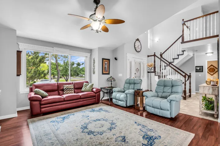 Wood-finish flooring, high ceilings, ceiling fan with integrated lighting, a prominent staircase with dark wood handrails, and large windows with decorative trim