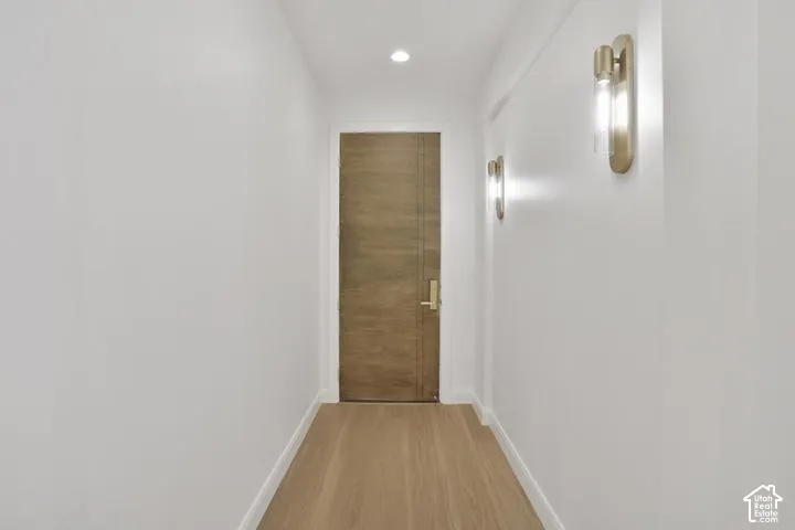 Wood-finish flooring, recessed ceiling lighting, and contemporary wall sconces illuminate this hallway leading to a solid panel door