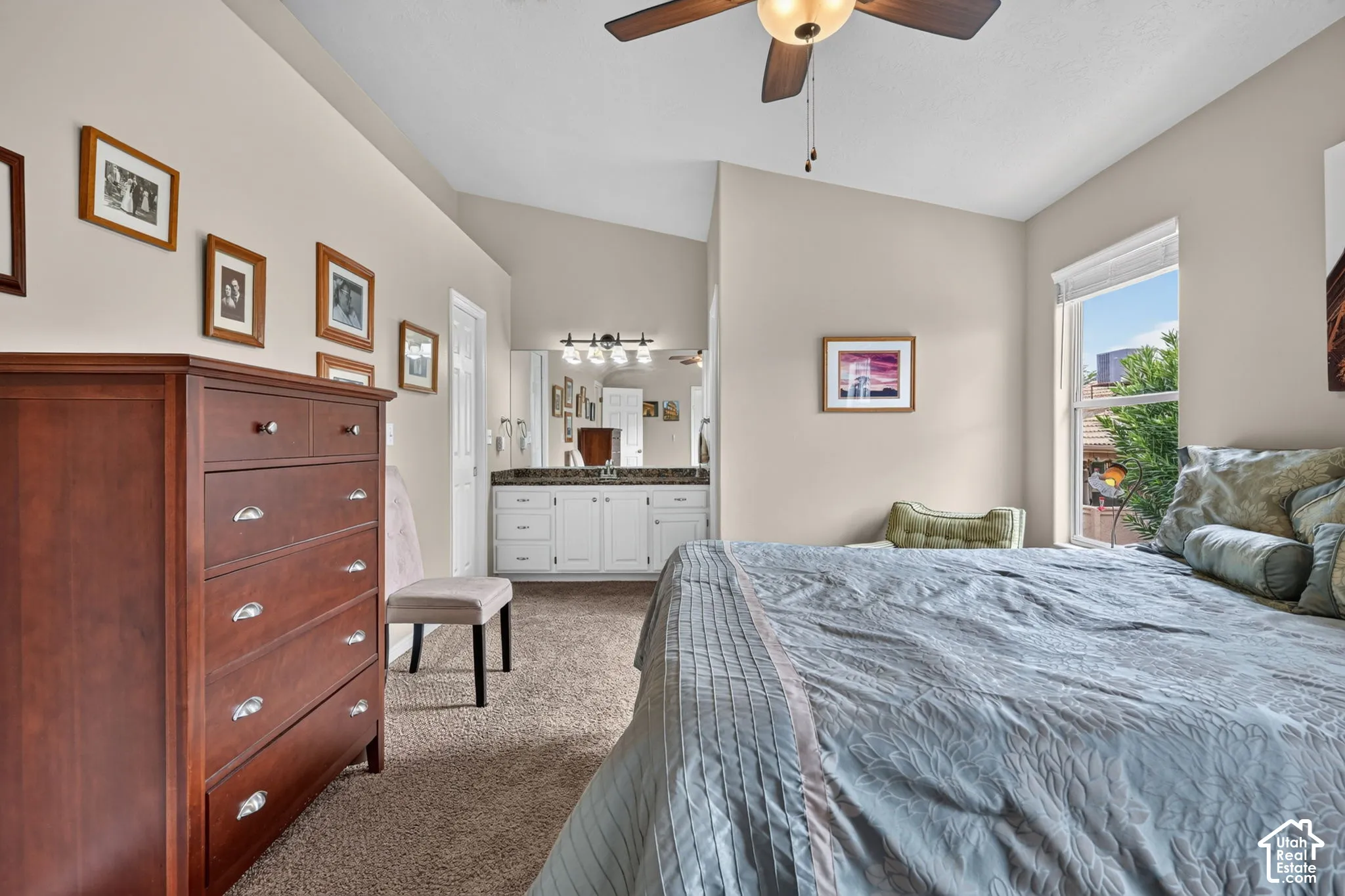 Vaulted ceiling with a ceiling fan, window with roller shade, integrated vanity with dual sinks, wall-mounted light fixture, and carpet flooring