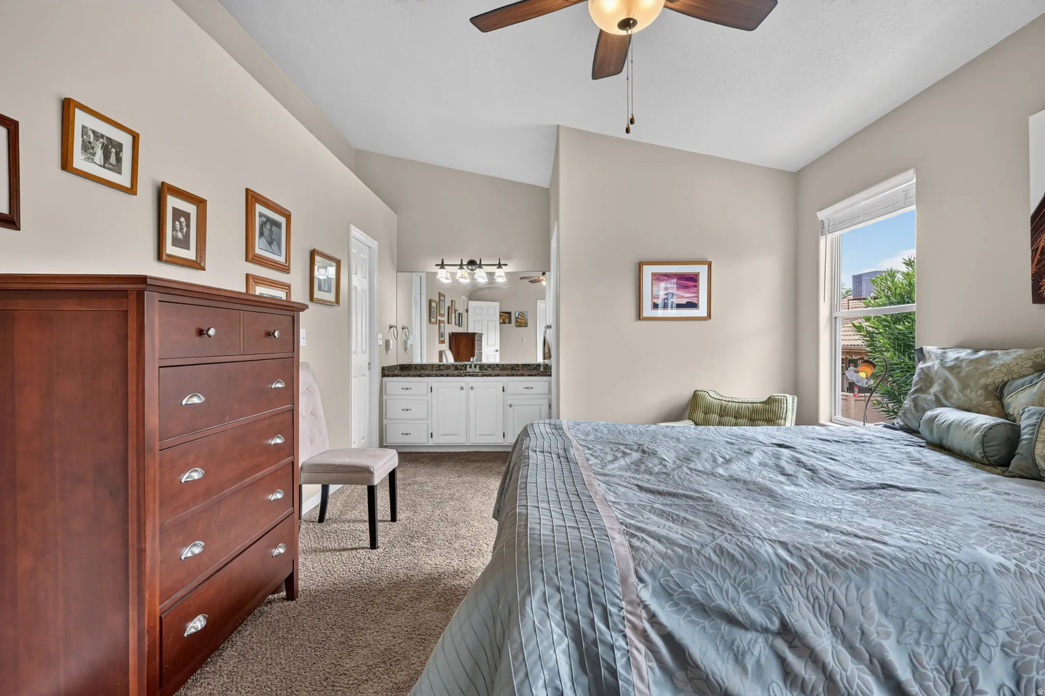 Vaulted ceiling with a ceiling fan, window with roller shade, integrated vanity with dual sinks, wall-mounted light fixture, and carpet flooring