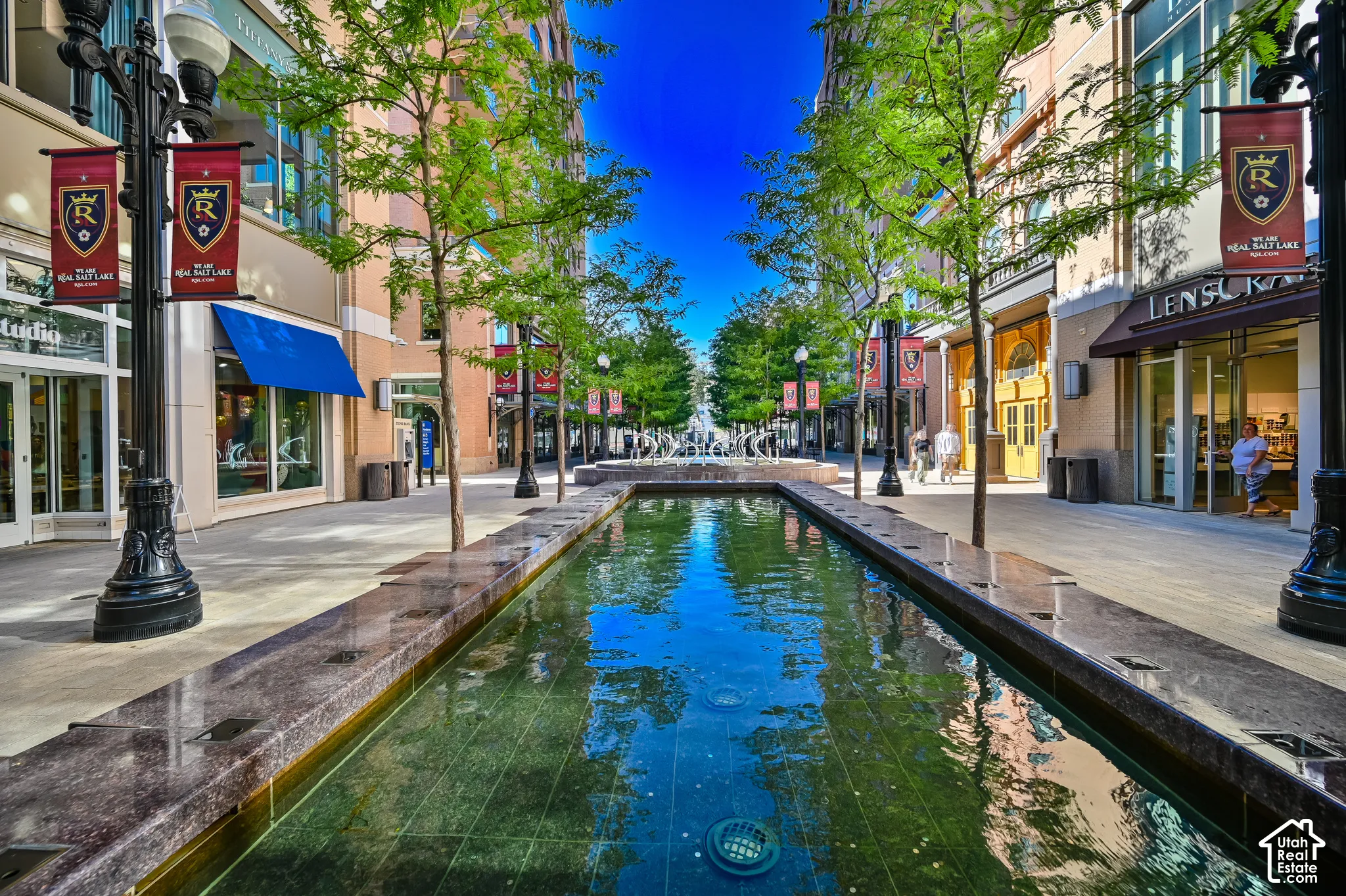 Urban plaza featuring a linear reflecting pool with stone edging, flanked by retail storefronts with large display windows, professional street lighting, mature tree canopy overhead, and paved pedestrian walkway