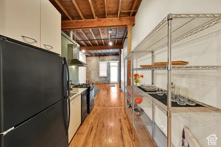 Galley-style kitchen featuring exposed wood-beam ceiling, black appliances, cream cabinetry, and wood-finish flooring throughout