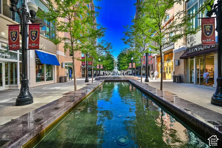 Urban plaza featuring a linear reflecting pool with stone edging, flanked by retail storefronts with large display windows, professional street lighting, mature tree canopy overhead, and paved pedestrian walkway