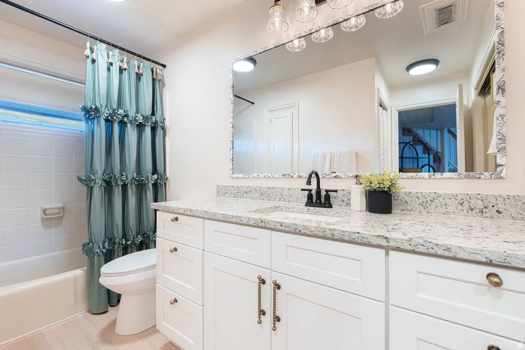 White shaker-style vanity with brass hardware, speckled countertop, undermount sink with matte black faucet, framed mirror, and tile surround bathtub