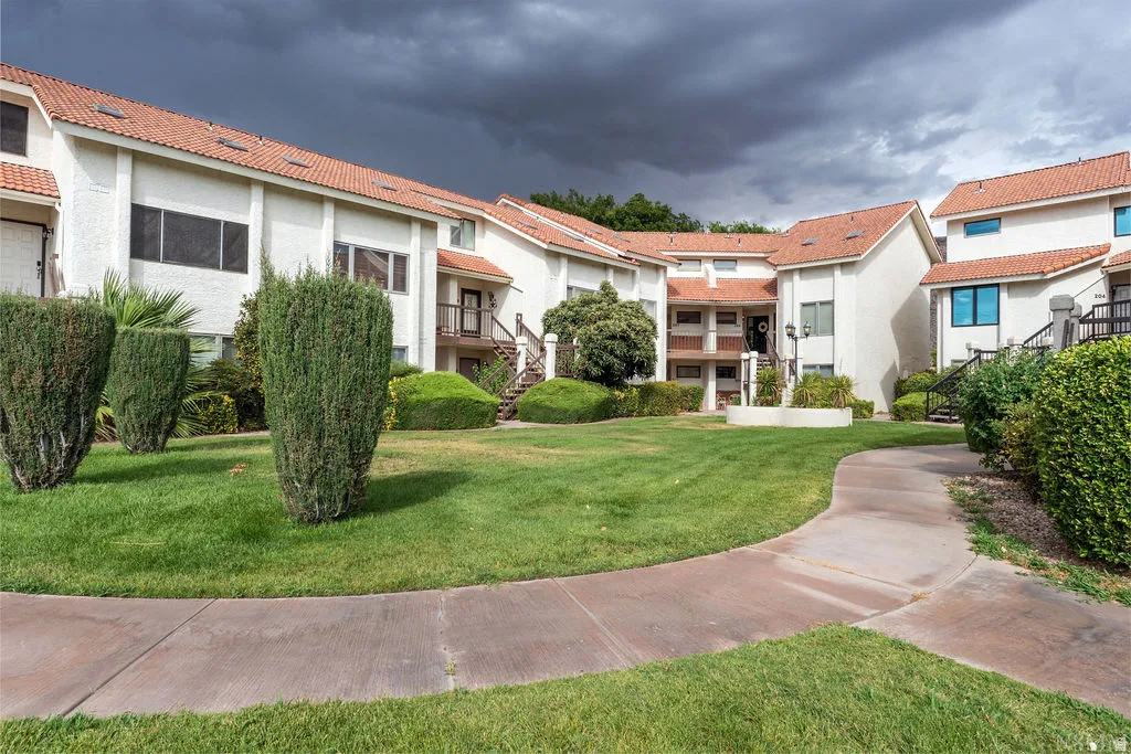 White stucco exterior with terracotta roof tiles, multiple balconies, a concrete pathway, and a manicured lawn with mature shrubs
