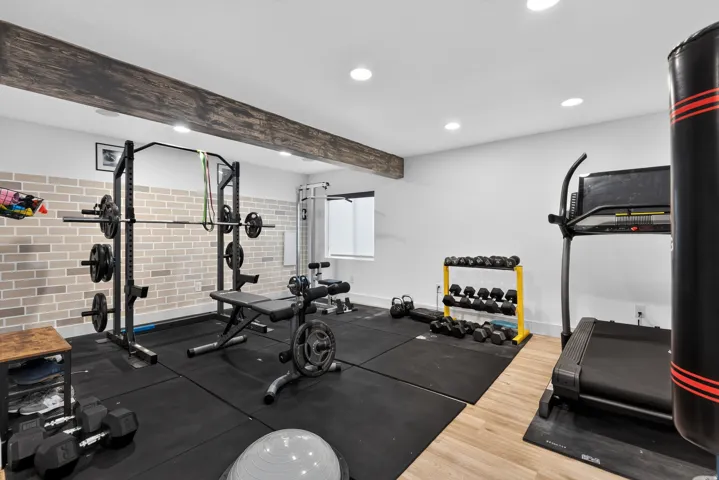 Recessed lighting illuminates the space, featuring a distressed wood-finish ceiling beam, a light-toned brick accent wall, and a combination of rubber mat and wood-finish flooring
