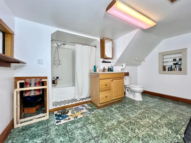 Green-patterned flooring, light wood-finish vanity with countertop, white toilet, and built-in shower with white tub