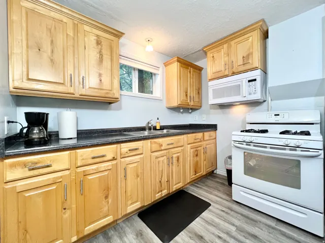 Wood-finish flooring, light wood cabinetry, white over-the-range microwave, white gas range, and a dual basin stainless steel sink