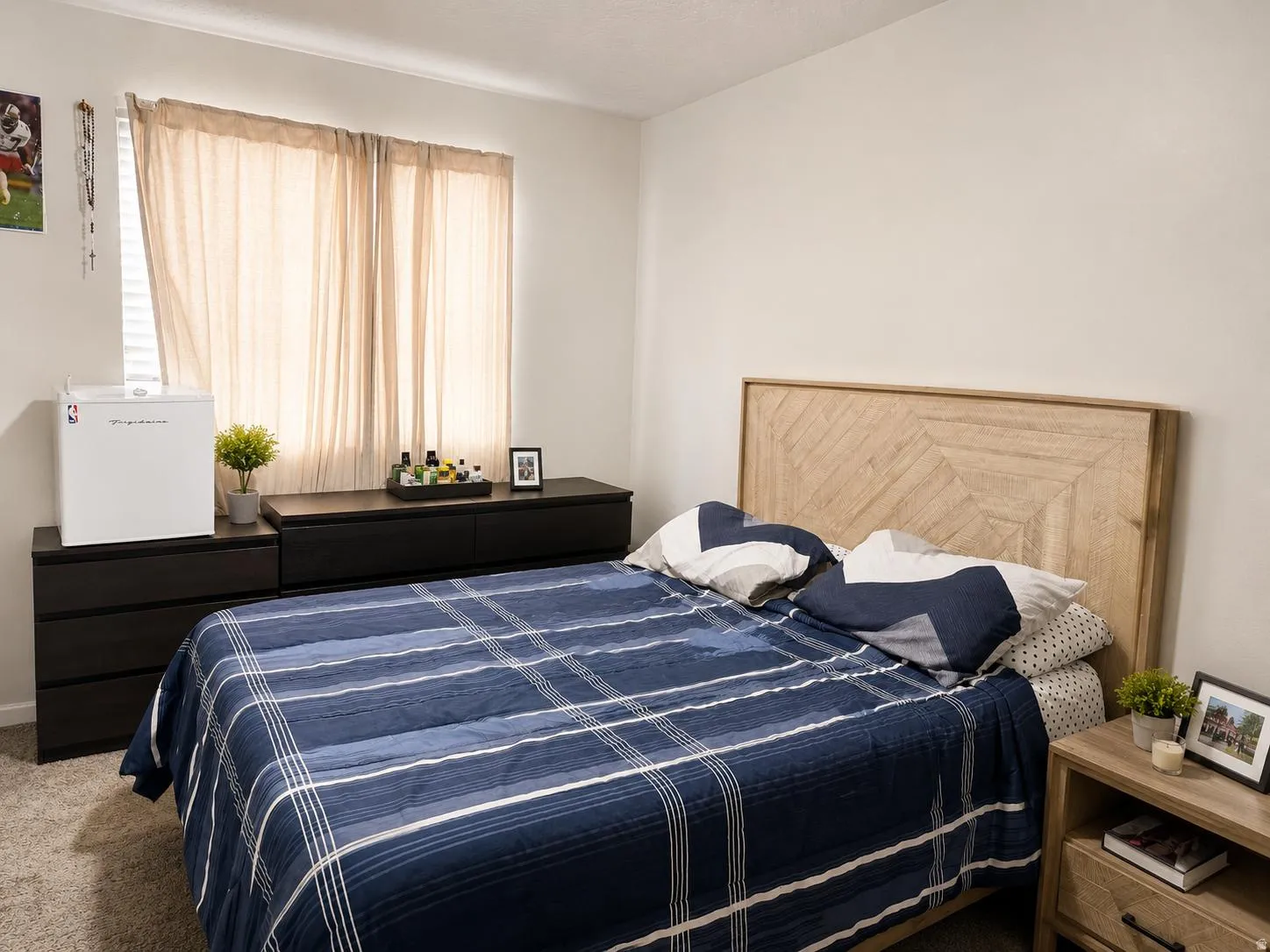 Light-colored walls, window with blinds, light-toned window coverings, headboard with geometric wood-finish pattern, carpeted flooring