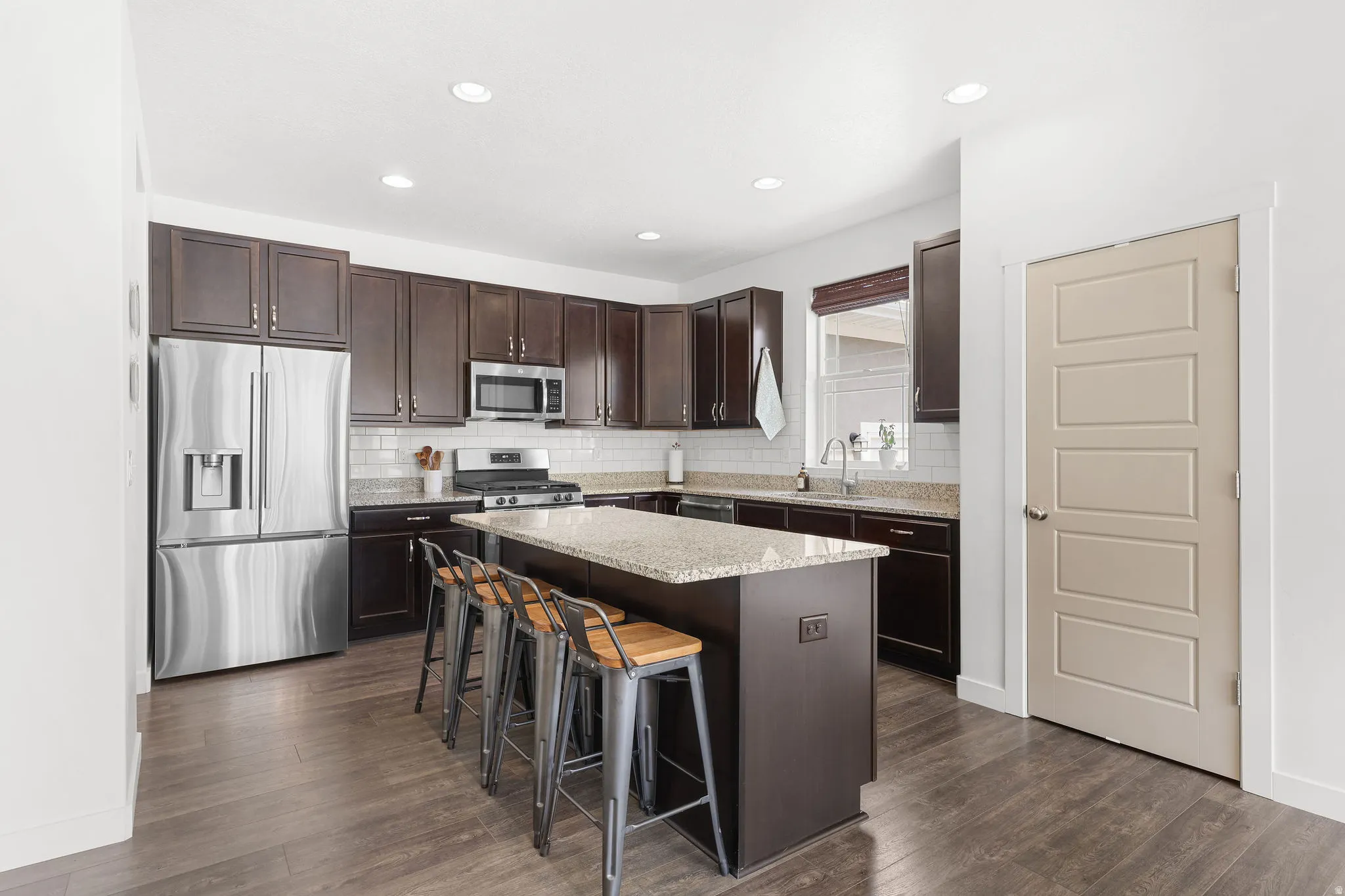 Kitchen featuring dark wood-finish cabinetry, stainless steel appliances, a light-toned granite island, a subway tile backsplash, and recessed lighting