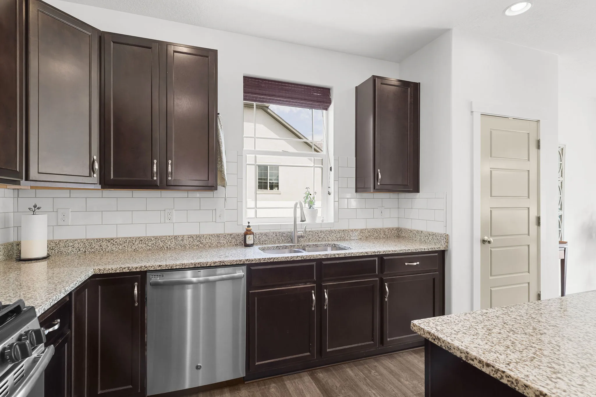 Dark wood-finish cabinetry, light-toned countertops, subway tile backsplash, stainless steel dishwasher, and a stainless steel sink with a gooseneck faucet