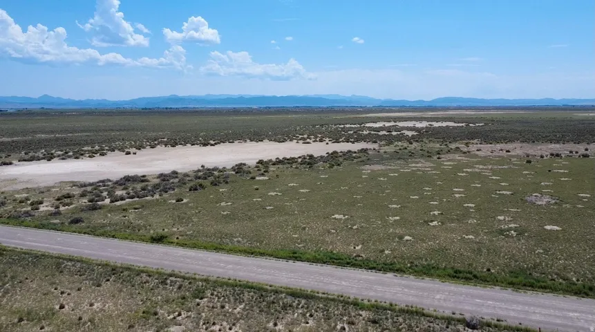 Expansive rural landscape with distant mountains and a clear sky, featuring a gravel road, open fields, and natural vegetation