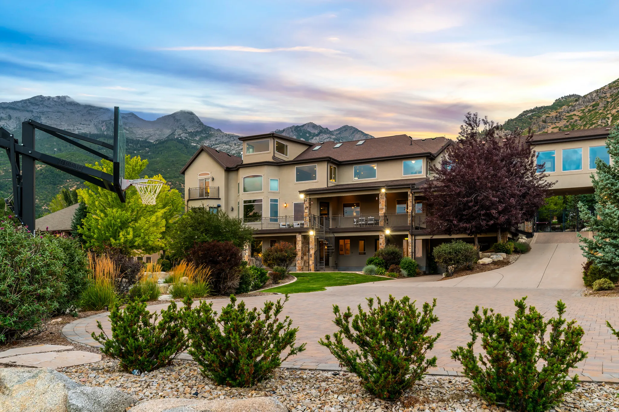 Multi-level stucco exterior with stone accents, featuring extensive landscaping, a paved driveway, and a prominent mountain backdrop