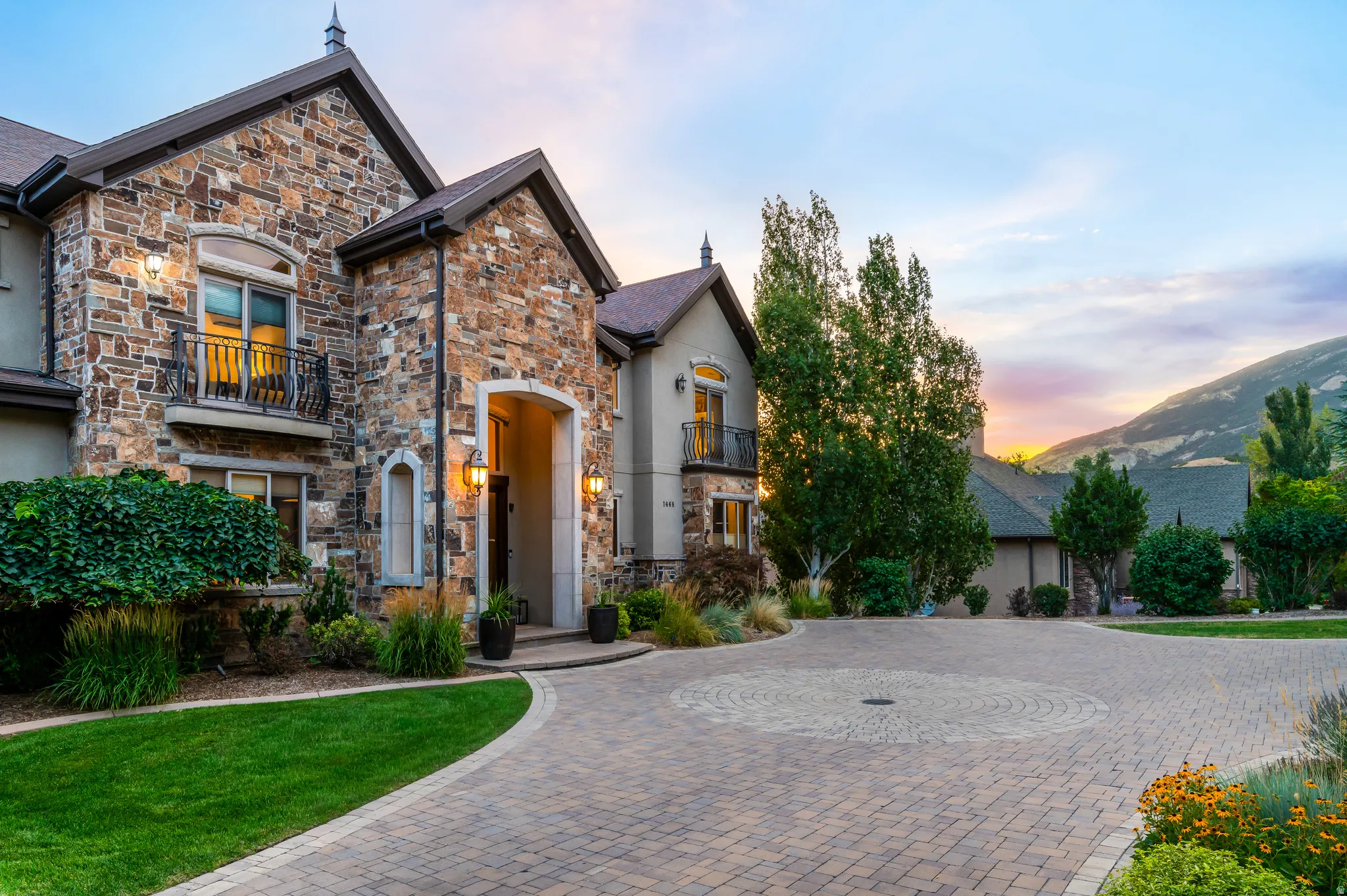 Stone facade exterior, arched entry, wrought iron balcony, paver driveway with circular pattern, and mature landscaping