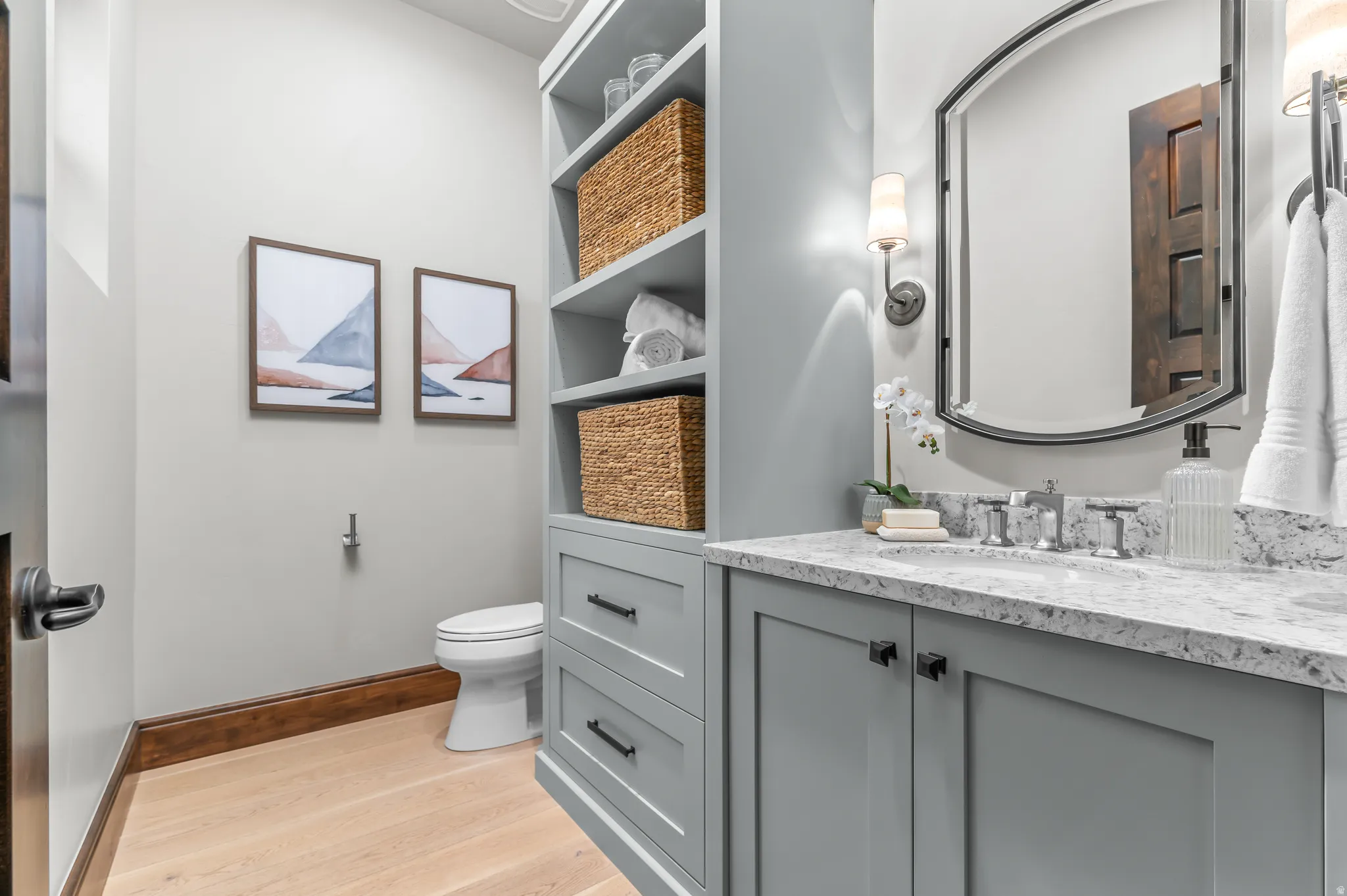 Light wood-finish flooring, gray vanity with stone countertop, built-in shelving, wall-mounted light fixtures, and a decorative mirror