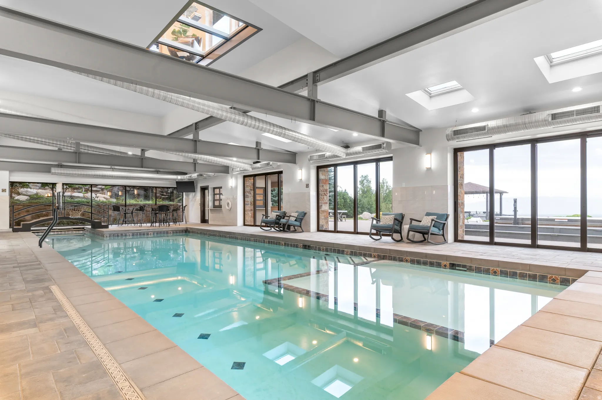 Indoor swimming pool with integrated spa, multiple skylights, full-height glass doors, and exposed ceiling beams