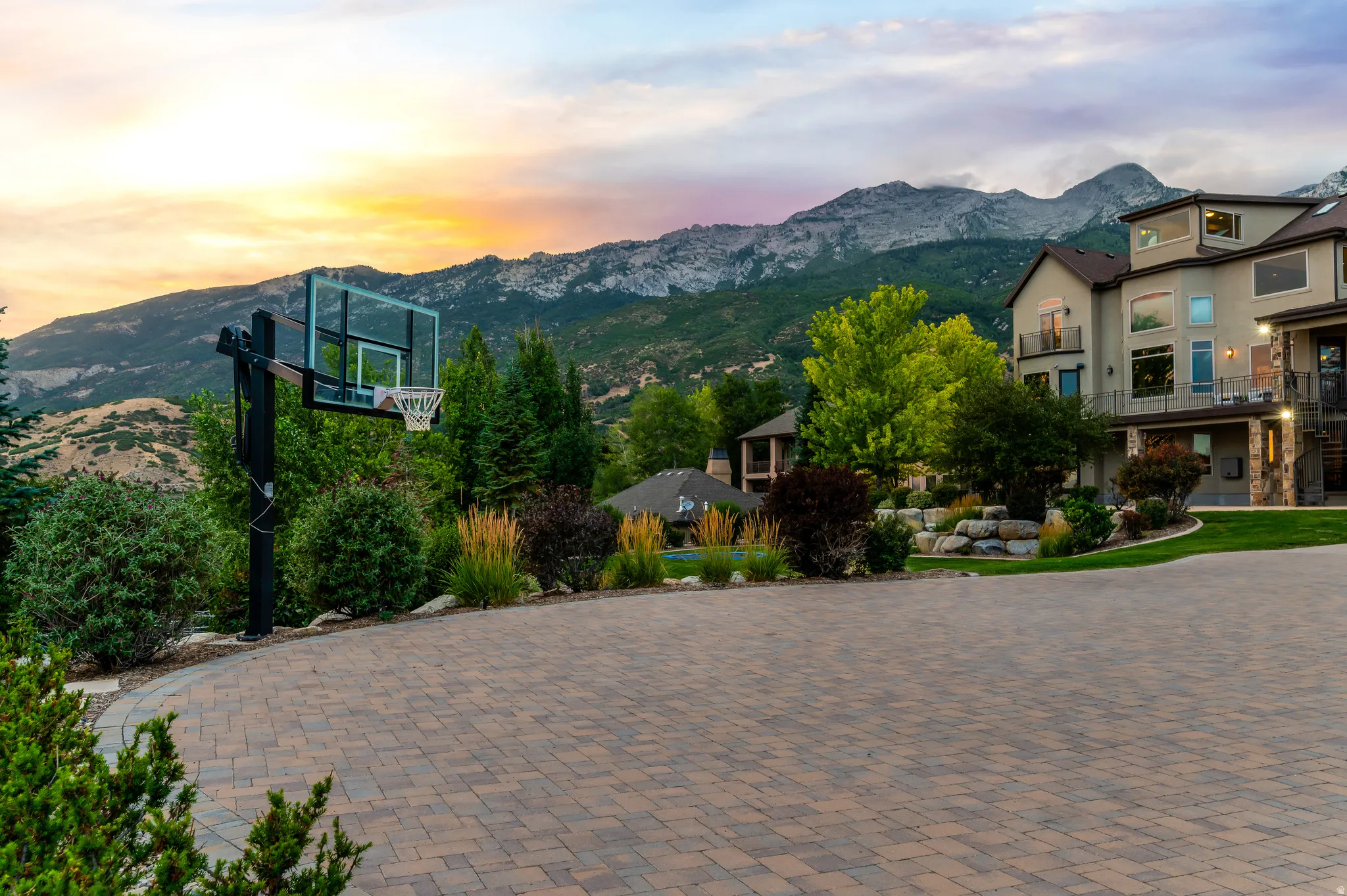 Paver driveway with a basketball hoop, multi-level exterior, stone accents, and mature landscaping