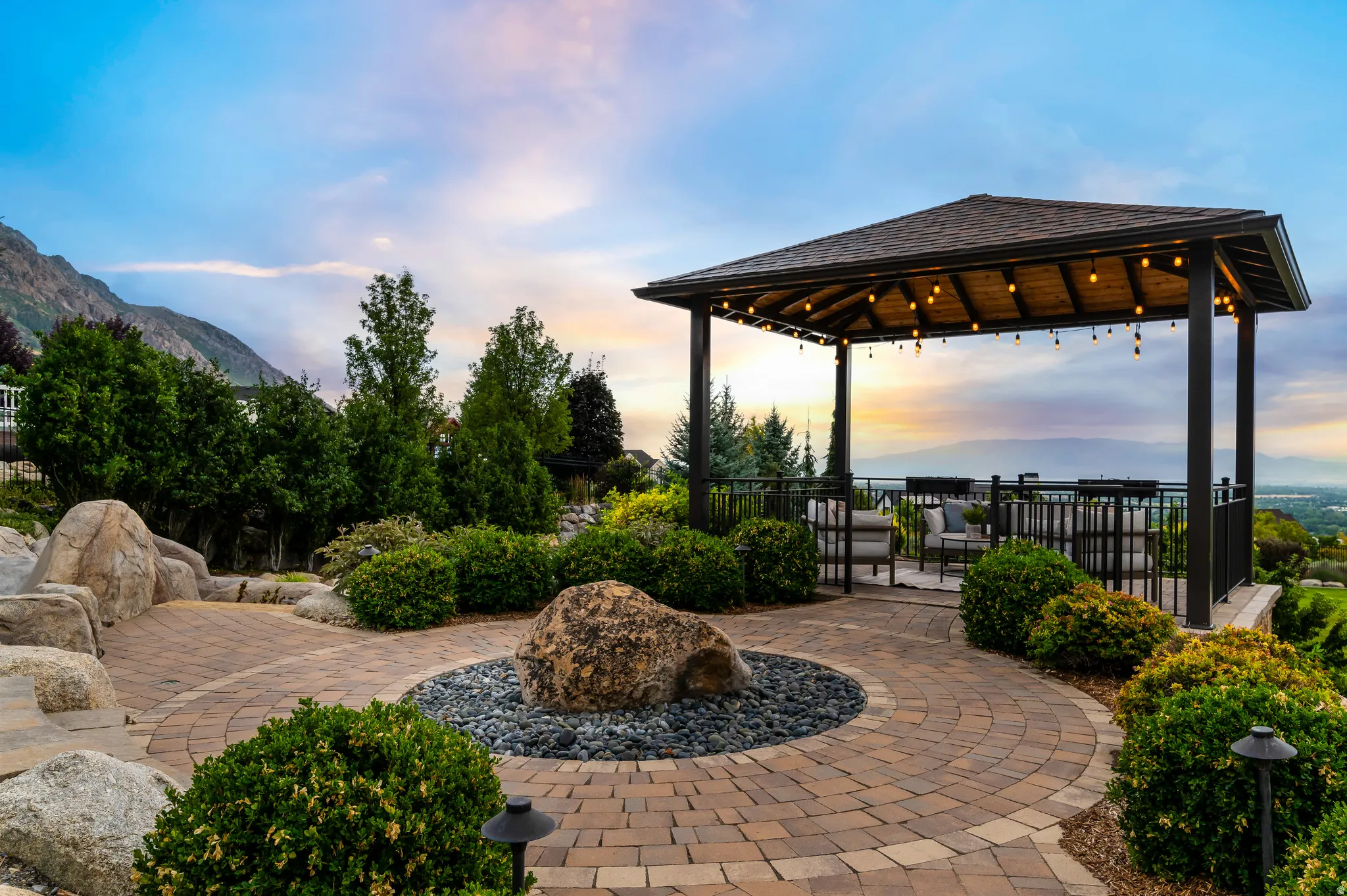 Expansive outdoor patio featuring a covered gazebo, paved walkways, mature landscaping, and mountain scenery
