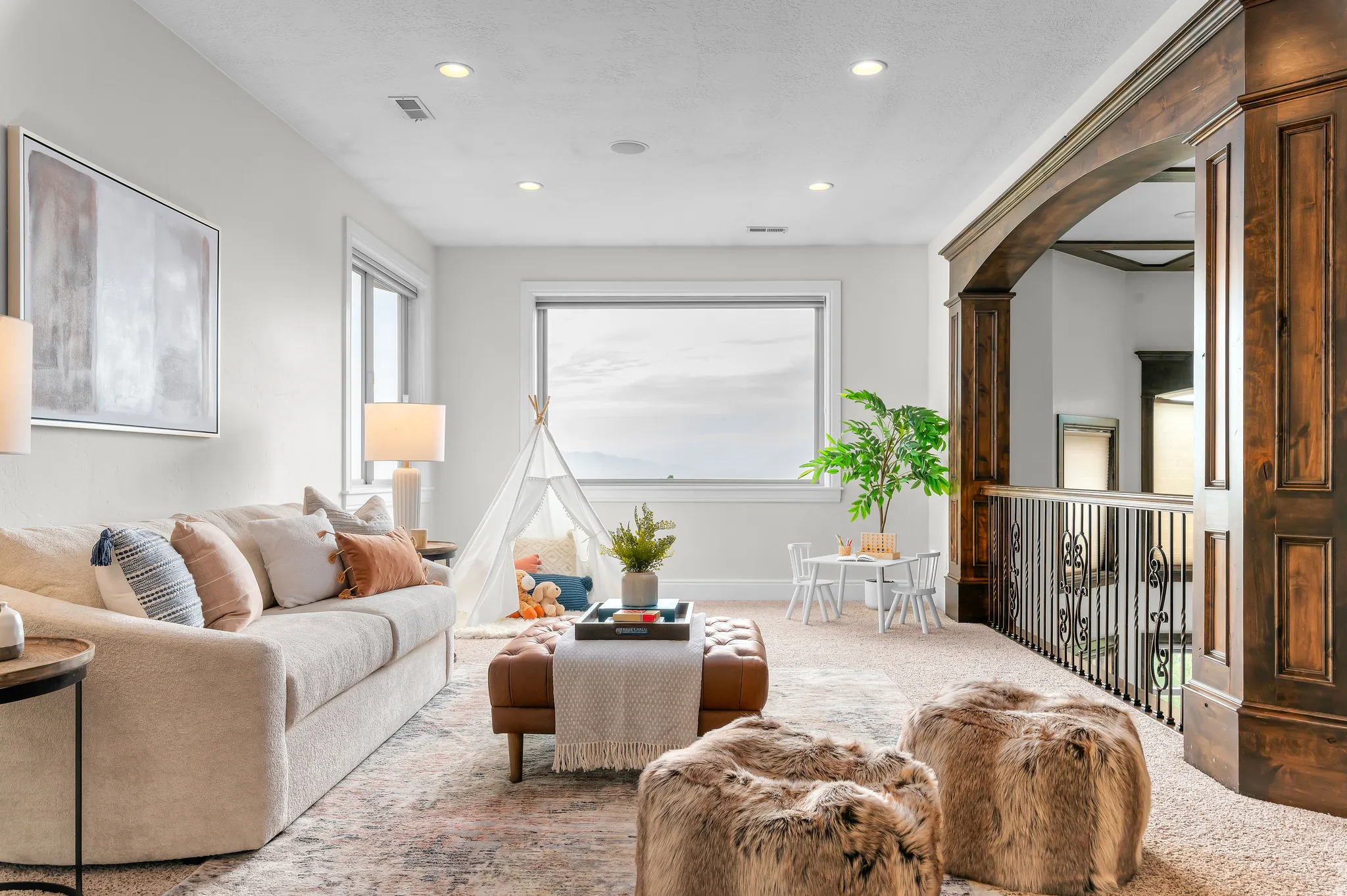 Recessed lighting illuminates a spacious room featuring a large window, warm-toned carpeting, and a dark wood architectural archway with wrought iron railing
