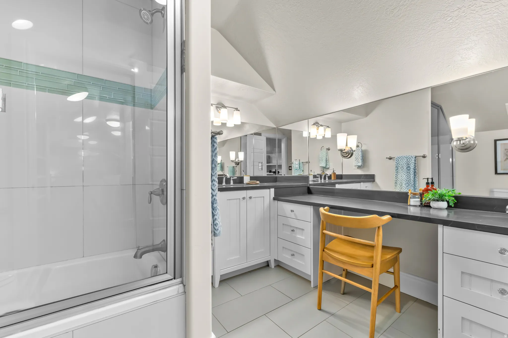 White cabinetry with brushed nickel hardware, expansive vanity with dark countertops, built-in vanity station, recessed lighting, and a glass-enclosed shower/tub combination with subway tile surround and decorative accent tiling