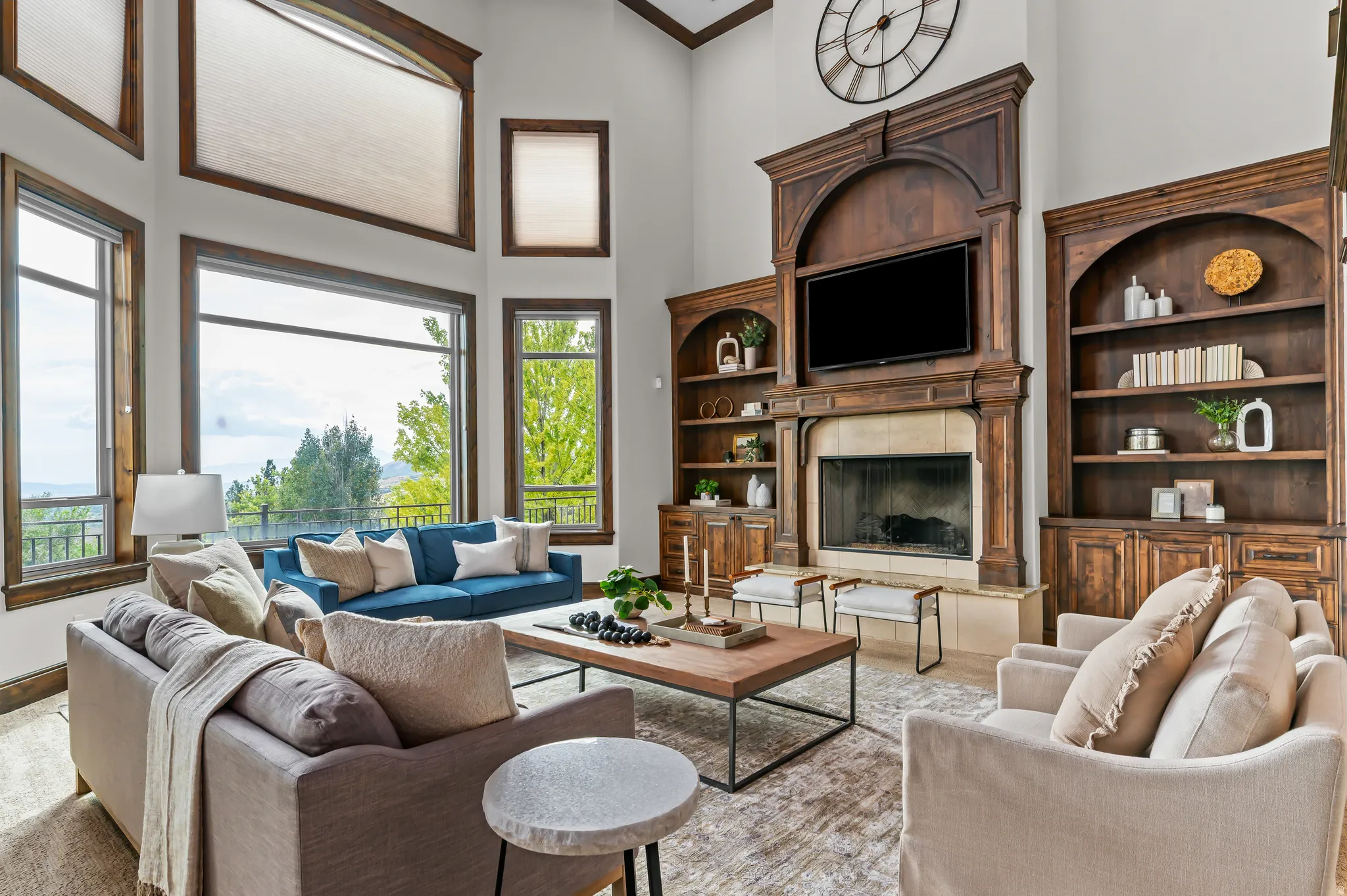 Two-story living area with high ceilings, large picture windows, a prominent wood-finish mantel and fireplace, and built-in wood-finish cabinetry