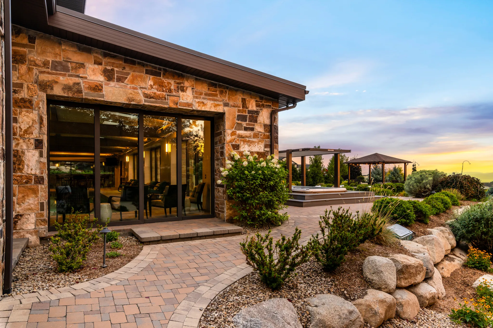 Stone-clad exterior featuring expansive glass doors, a paver patio, integrated hot tub with pergola, and a tiered rock garden with mature landscaping